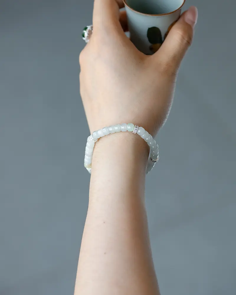 DSC_0679 拷贝 A hand wearing a delicate white beaded bracelet holds a small cup. The person also has a green gemstone ring on their index finger. The background is plain and gray.