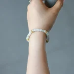 A hand wearing a beaded bracelet with white and gold accents holds a small, light blue ceramic cup against a plain, neutral background.