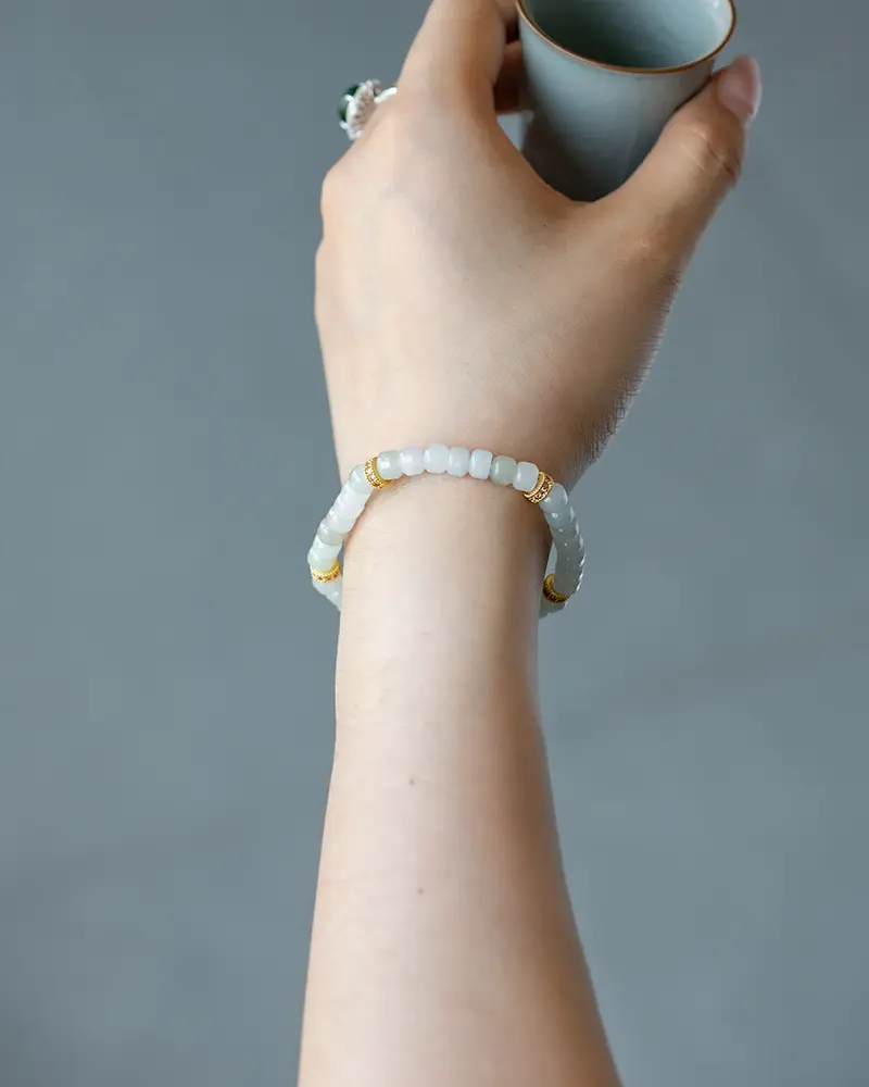 DSC_0681 拷贝 A hand wearing a beaded bracelet with white and gold accents holds a small, light blue ceramic cup against a plain, neutral background.