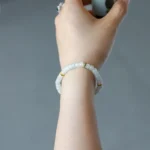 A hand holding a small cup, wearing a bracelet made of white beads with gold accents, against a plain gray background.