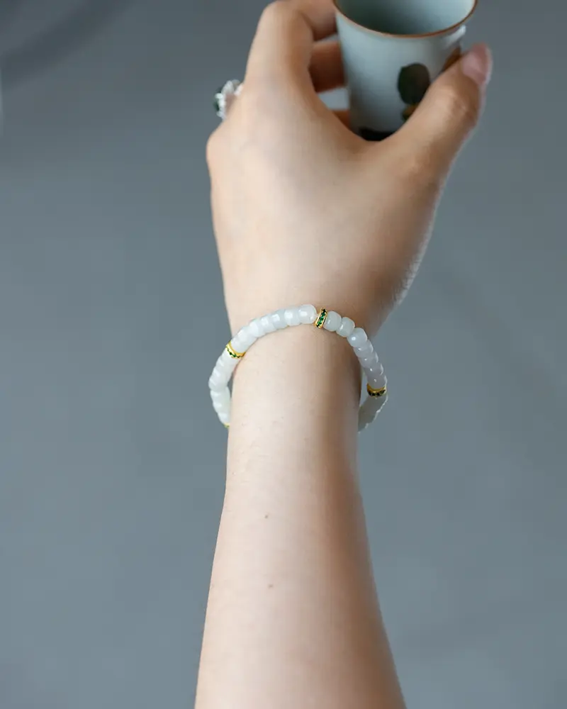 DSC_0683 拷贝 A hand holding a small cup, wearing a bracelet made of white beads with gold accents, against a plain gray background.