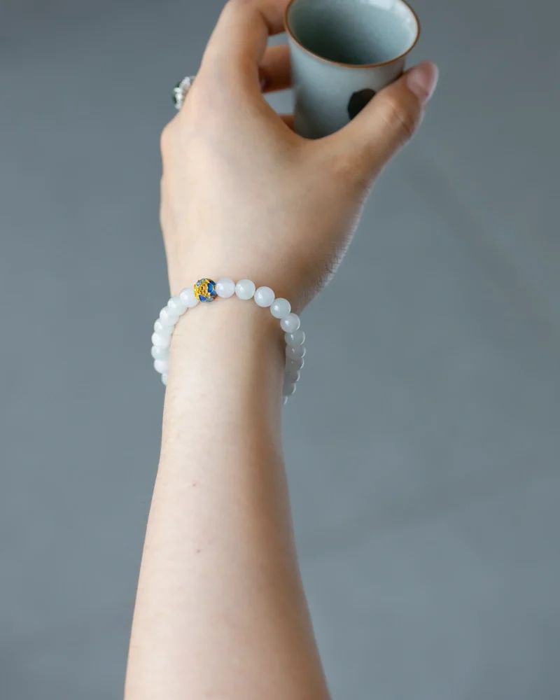 A hand wearing the White and Light Green Jadeite Bead Bracelet, featuring spherical beads with a blue accent detail, is holding a small ceramic cup against a plain gray background.