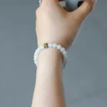 A hand holding a small cup is shown wearing a white beaded bracelet with a single yellow and blue accent bead, against a plain gray background.