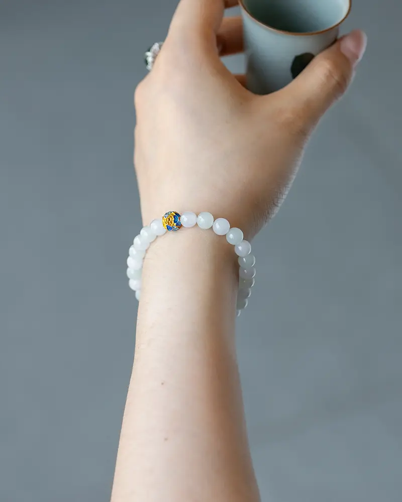 DSC_0688 拷贝 A hand holding a small cup is shown wearing a white beaded bracelet with a single yellow and blue accent bead, against a plain gray background.