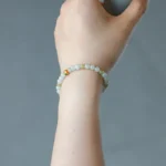 A hand holding a cup, wearing a delicate beaded bracelet with pale green and small orange and gold accent beads, against a plain gray background.