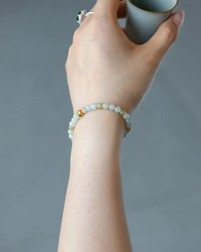 DSC_0692 拷贝 A hand holding a cup, wearing a delicate beaded bracelet with pale green and small orange and gold accent beads, against a plain gray background.
