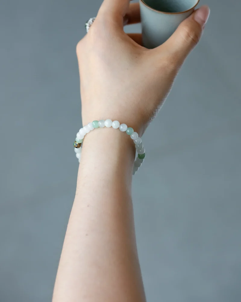 A hand holding a cup and wearing the Light Green and White Jadeite Bead Bracelet — featuring spherical beads with an enamel-style accent—set against a blurred gray background.