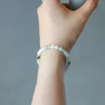 A hand holding a cylindrical object, wearing a beaded bracelet with light green and white round stones. The background is plain and out of focus.