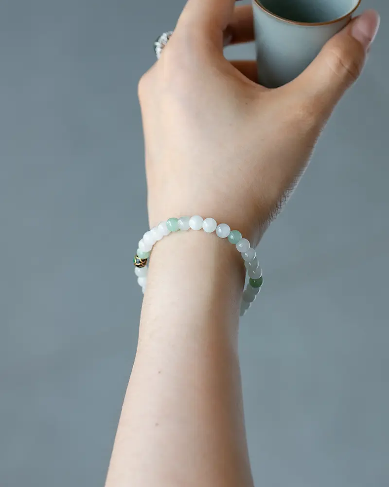DSC_0693 拷贝 A hand holding a cylindrical object, wearing a beaded bracelet with light green and white round stones. The background is plain and out of focus.