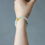 A hand wearing a beaded bracelet made of pale green stones with a single gold-colored bead, shown against a plain gray background.
