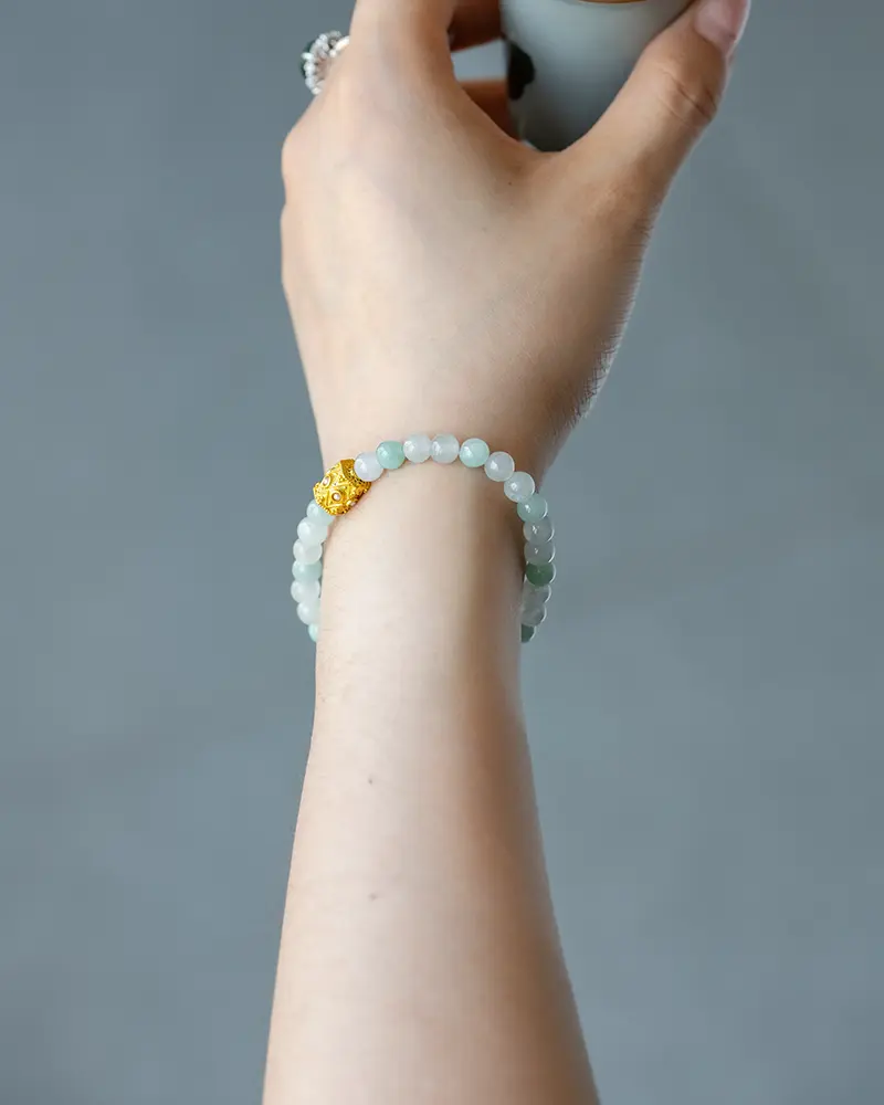 DSC_0694 拷贝 A hand wearing a beaded bracelet made of pale green stones with a single gold-colored bead, shown against a plain gray background.