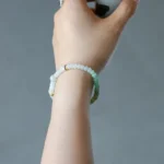 A hand wearing a beaded bracelet in shades of white and green holds a small white cup against a plain gray background.