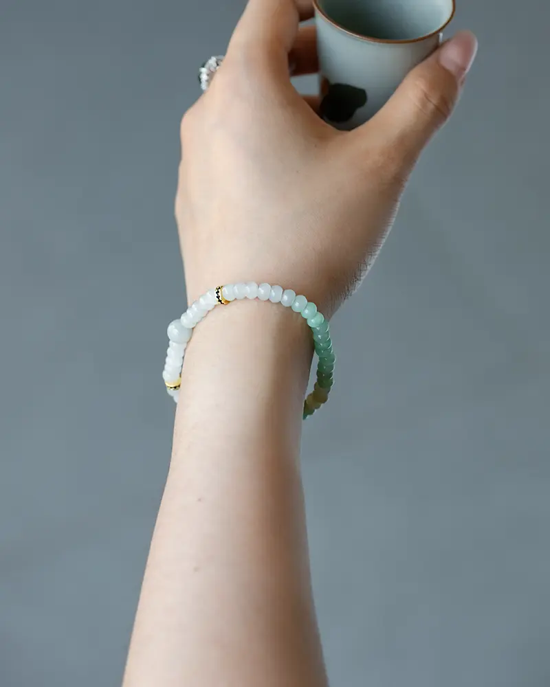 A hand wearing a beaded bracelet in shades of white and green holds a small white cup against a plain gray background.