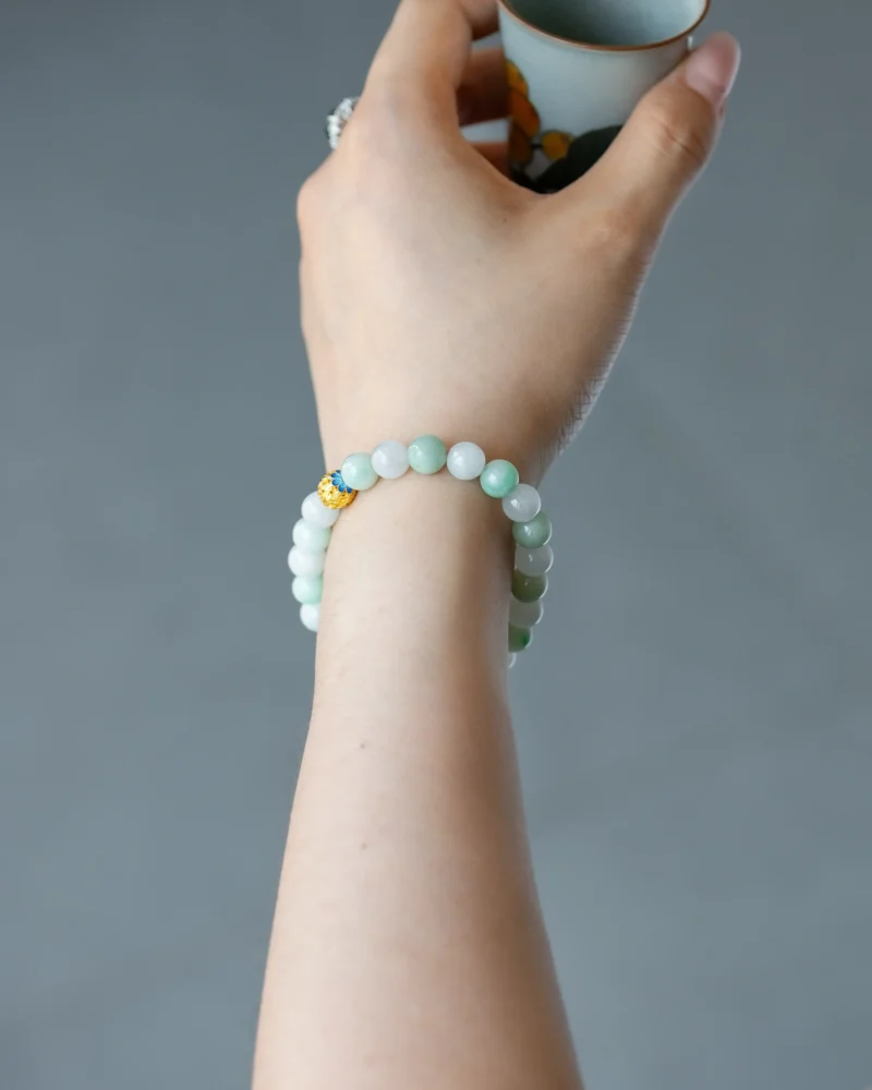 A hand holding a small cup, adorned with the Light Green and White Jadeite Bead Bracelet—spherical beads in light green and white jade, finished with a gold-tone accent. The background is plain and gray.