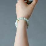 A hand wearing a double-strand light green beaded bracelet with one yellow bead holds a small cup, against a plain grey background.