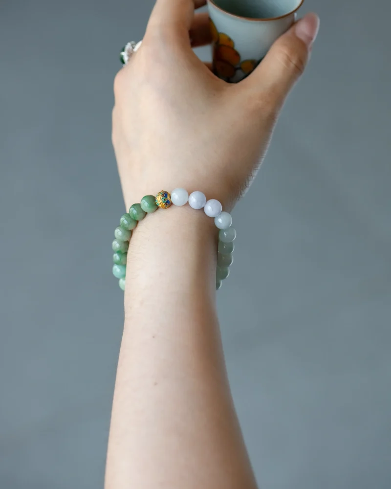 A hand wearing the Light Green, Light Purple, and Brownish Yellow Jadeite Bead Bracelet with spherical beads and a gold-tone accent detail holds a small cup against a blurred gray background.
