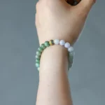 A hand wearing a beaded bracelet made of green and white round stones with one gold and blue accent bead, against a neutral gray background.