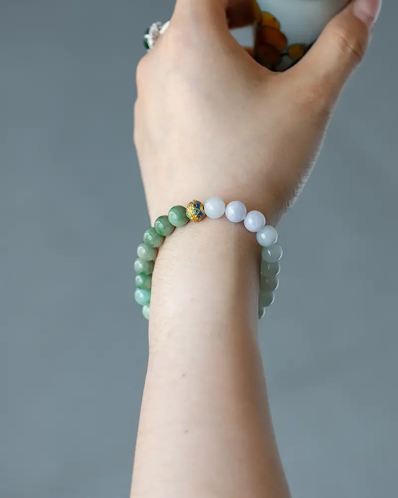 DSC_0698 拷贝 A hand wearing a beaded bracelet made of green and white round stones with one gold and blue accent bead, against a neutral gray background.