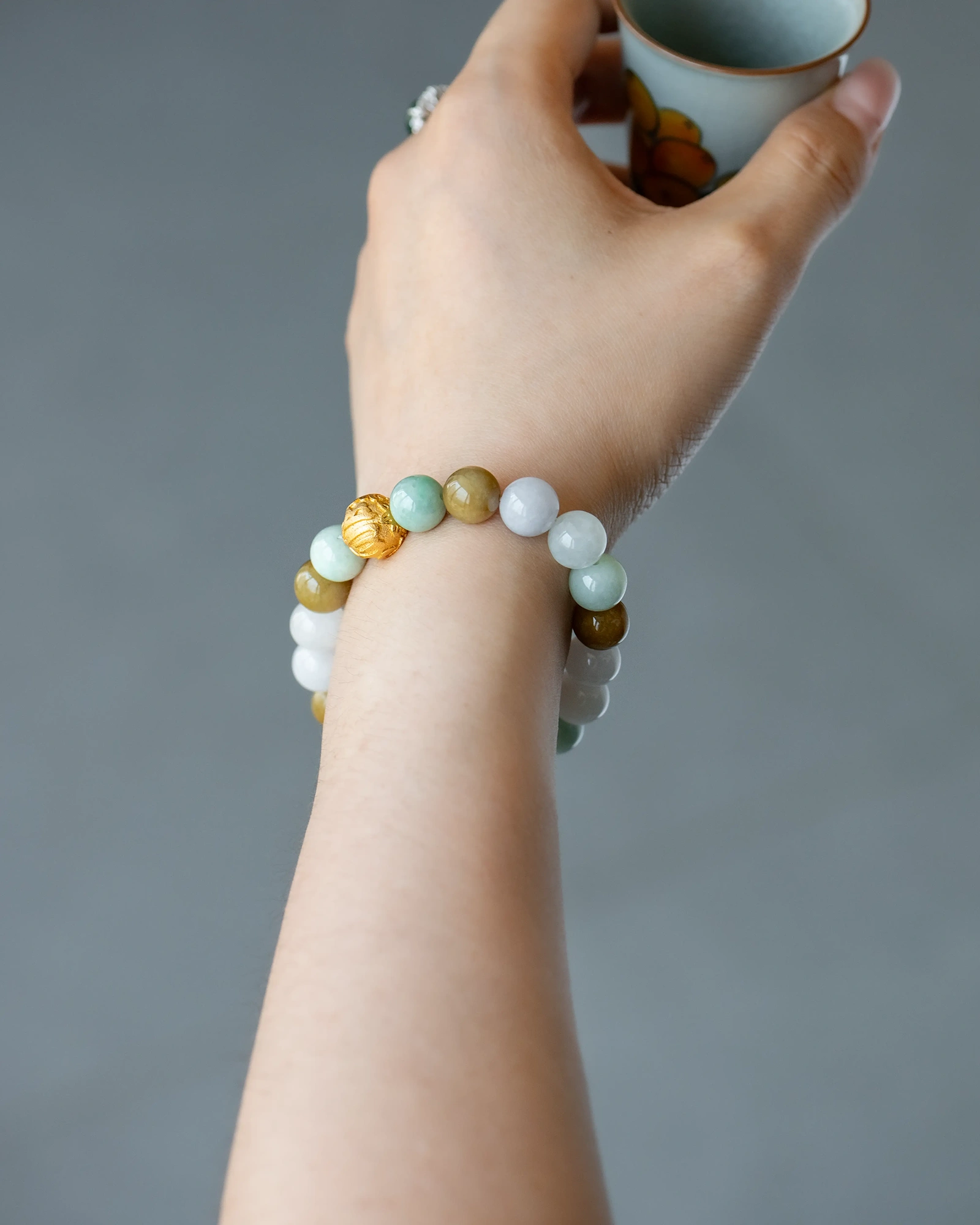 A hand holding a small cup, wearing the Light Green, White, and Brownish Yellow Jadeite Bead Bracelet featuring spherical beads with gold-tone accents, against a plain gray background.