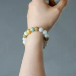 A hand holding a small cup, wearing a beaded bracelet made of green, white, and yellow round stones with one gold bead. The background is plain gray.