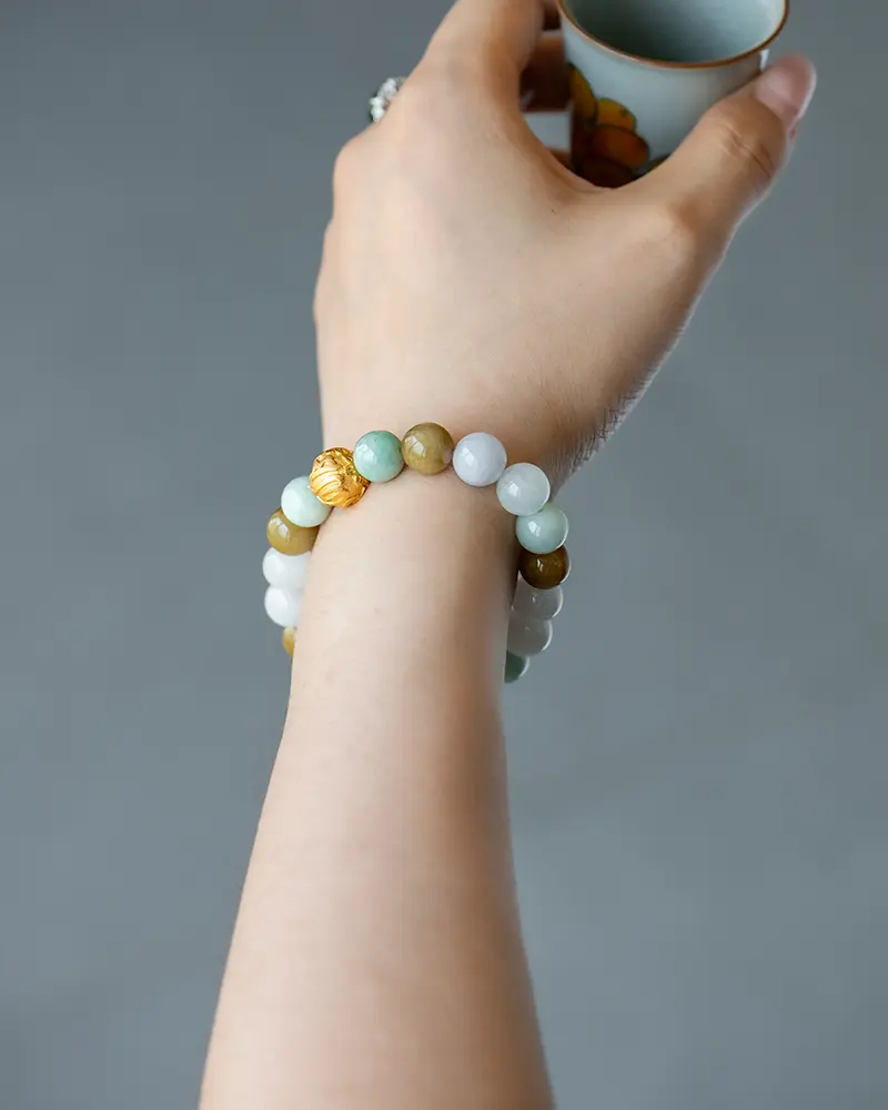DSC_0699 拷贝 A hand holding a small cup, wearing a beaded bracelet made of green, white, and yellow round stones with one gold bead. The background is plain gray.