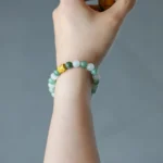 A hand wearing a green and white beaded bracelet with a gold accent holds a small cup decorated with a yellow-orange floral design. The background is blurred gray.