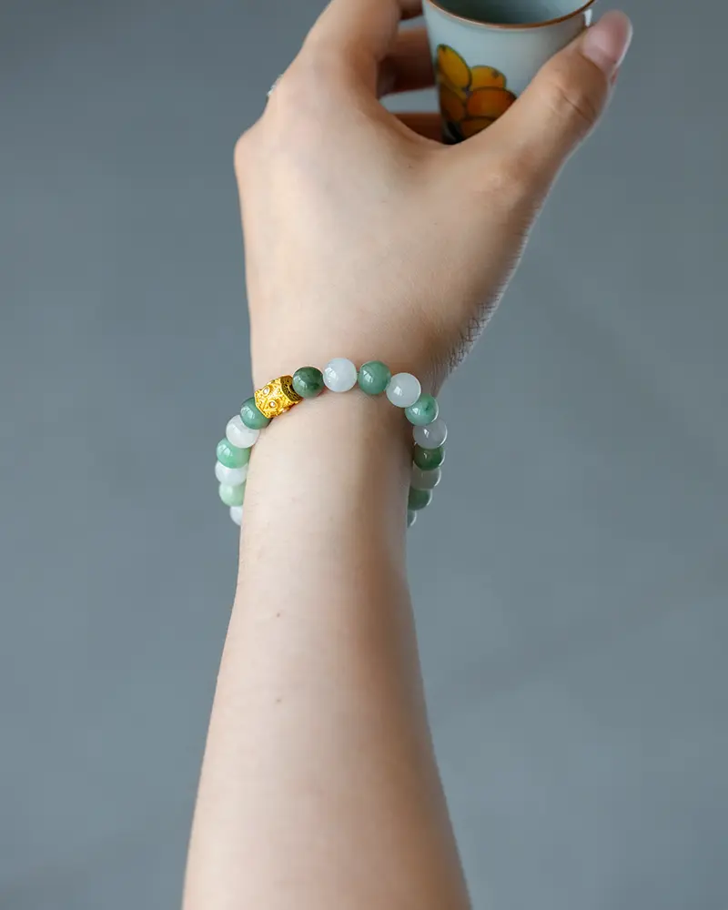DSC_0701 拷贝 2 A hand wearing a green and white beaded bracelet with a gold accent holds a small cup decorated with a yellow-orange floral design. The background is blurred gray.