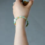 A hand holding a small cup is shown. The person is wearing two beaded bracelets in green, white, and orange tones on their wrist. The background is plain and gray.