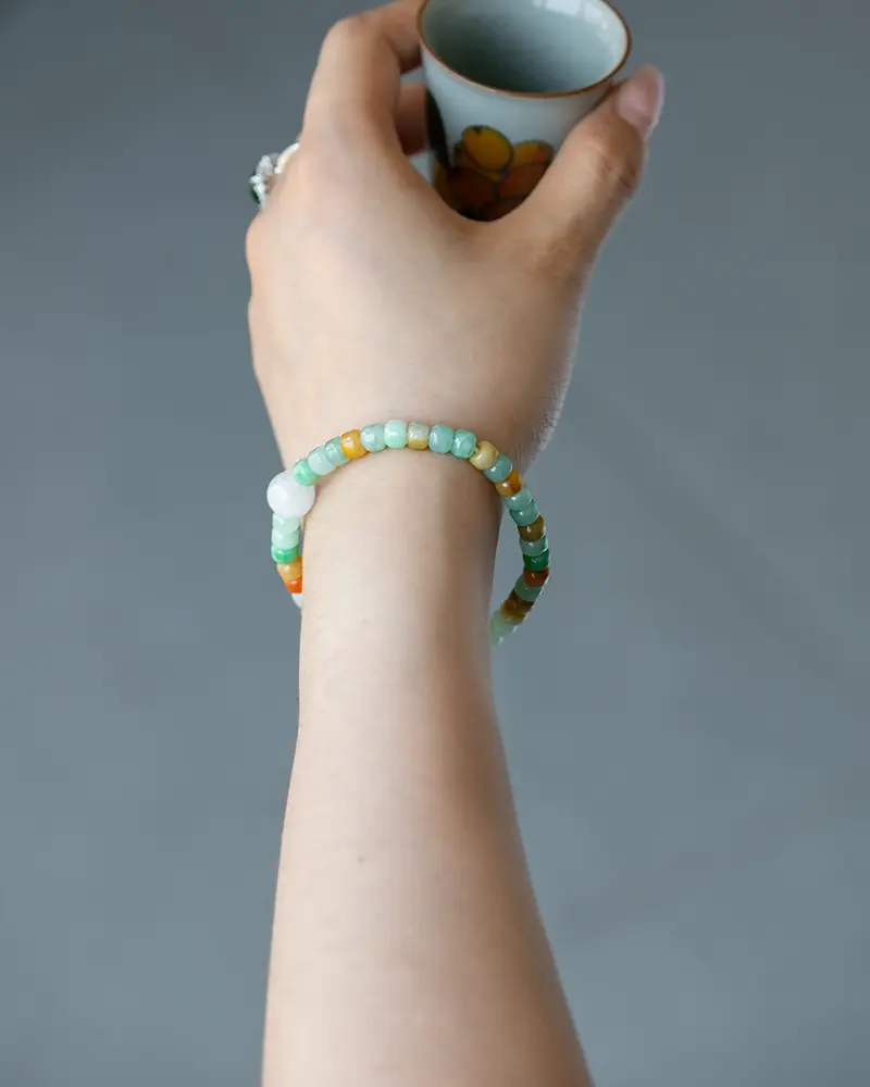 DSC_0703 拷贝 A hand holding a small cup is shown. The person is wearing two beaded bracelets in green, white, and orange tones on their wrist. The background is plain and gray.