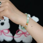 A person wearing a colorful beaded bracelet and a silver ring on their hand, with a black shirt featuring white bear illustrations with pink bows in the background.