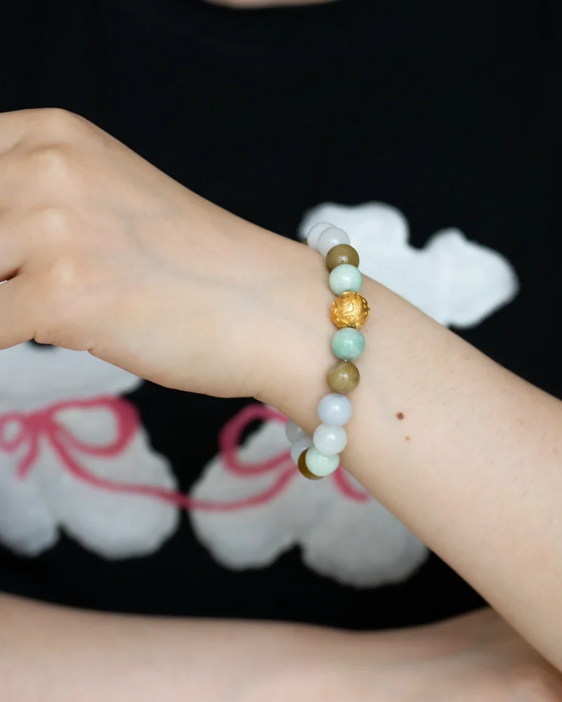 A person wearing the Light Green, White, and Brownish Yellow Jadeite Bead Bracelet—featuring spherical beads with a gold-tone accent—against a black shirt with a white and pink pattern.