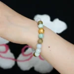 A close-up of a wrist wearing a beaded bracelet with light green, white, brown, and a gold textured bead, set against a black shirt with white and pink designs in the background.