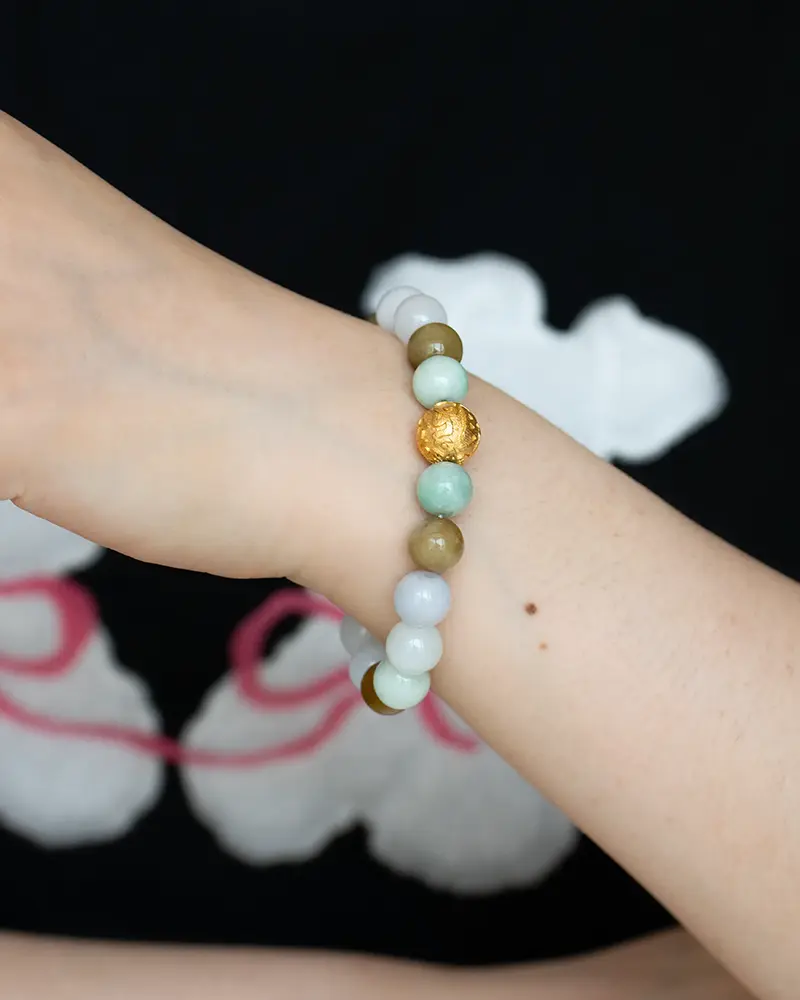 DSC_0705 拷贝 A close-up of a wrist wearing a beaded bracelet with light green, white, brown, and a gold textured bead, set against a black shirt with white and pink designs in the background.