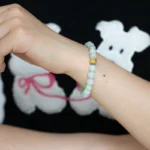 A person wearing a light green beaded bracelet with a single gold bead, shown against a black shirt featuring a white cartoon dog with a pink leash.