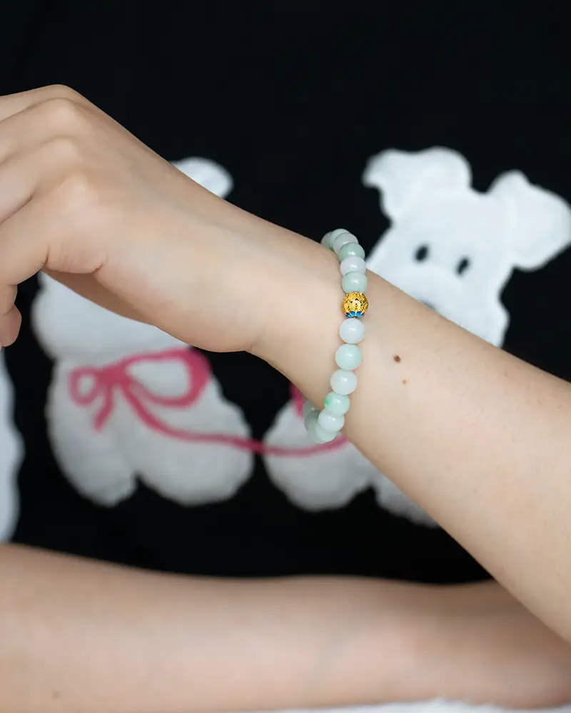 A person wearing a light green beaded bracelet with a single gold bead, shown against a black shirt featuring a white cartoon dog with a pink leash.