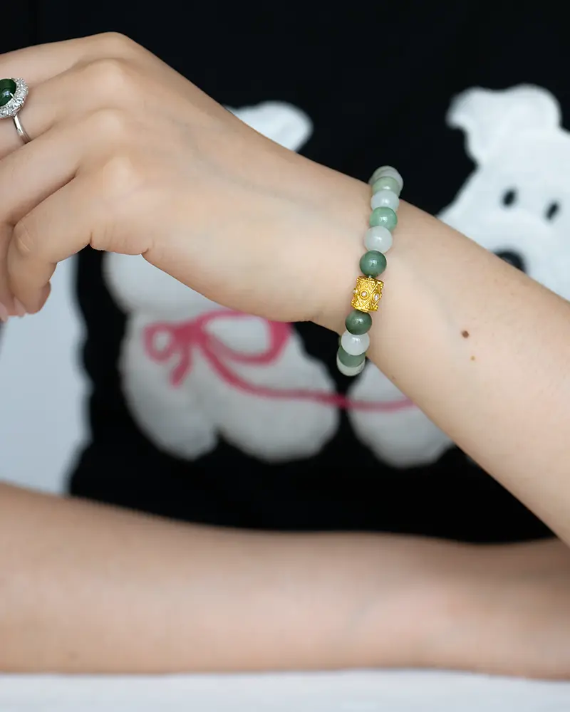 DSC_0708 拷贝 A hand wearing a green beaded bracelet with a gold charm rests on a table. The person also wears a green gemstone ring, and has a shirt featuring a cartoon bear with a pink ribbon.
