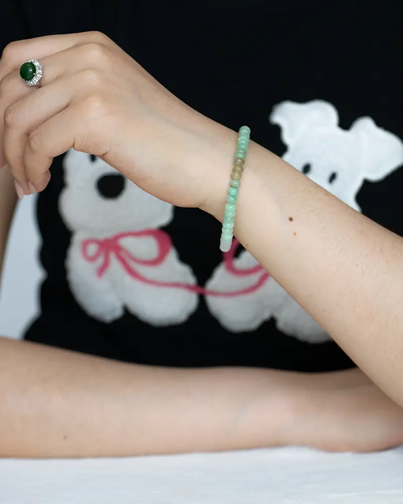 A person wearing a beaded green bracelet and a green gemstone ring rests their arm on a table. They are wearing a black shirt with a graphic of two white dogs with red leashes.