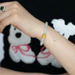 A person wearing a beaded bracelet with a gold accent on their wrist and a green stone ring on their finger. The person is wearing a black shirt with white cartoon bears and pink details.