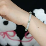 A hand wearing a beaded bracelet with light green and white beads is raised against a black background featuring a teddy bear design with pink bows.