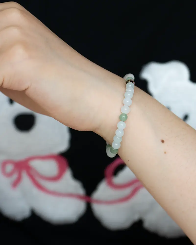 DSC_0719 拷贝 A hand wearing a beaded bracelet with light green and white beads is raised against a black background featuring a teddy bear design with pink bows.