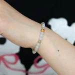 A close-up of a wrist wearing a beaded bracelet with light-colored round beads and one bead accented in gold and red, against a dark background with white and pink abstract shapes.