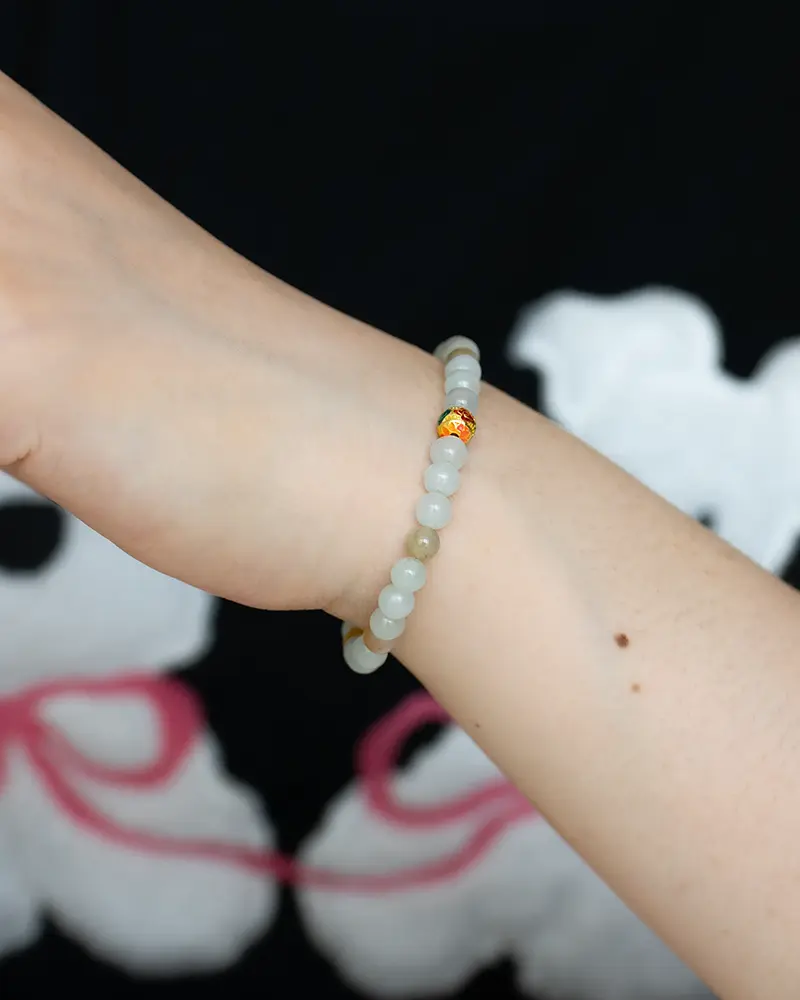 DSC_0720 拷贝 A close-up of a wrist wearing a beaded bracelet with light-colored round beads and one bead accented in gold and red, against a dark background with white and pink abstract shapes.