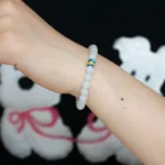 A hand wearing a white beaded bracelet with a single blue and yellow bead, in front of a black shirt featuring a white cartoon bear design with pink accents.