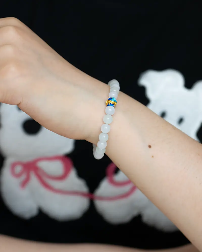 DSC_0721 拷贝 A hand wearing a white beaded bracelet with a single blue and yellow bead, in front of a black shirt featuring a white cartoon bear design with pink accents.