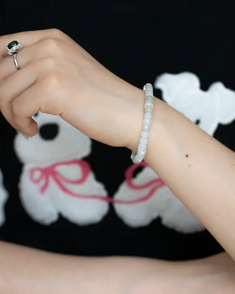 DSC_0723 拷贝 A hand wearing a silver ring and a beaded white bracelet is posed in front of a black shirt with a white teddy bear graphic and pink ribbon accents.