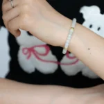 A person wearing a beaded bracelet and a ring rests their arm on a surface. The person is wearing a black shirt with a white bear graphic holding red string.