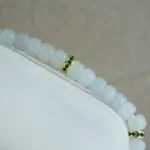A close-up of a beaded bracelet featuring translucent white beads and two gold spacer beads accented with small green stones, displayed on a white surface with a neutral background.