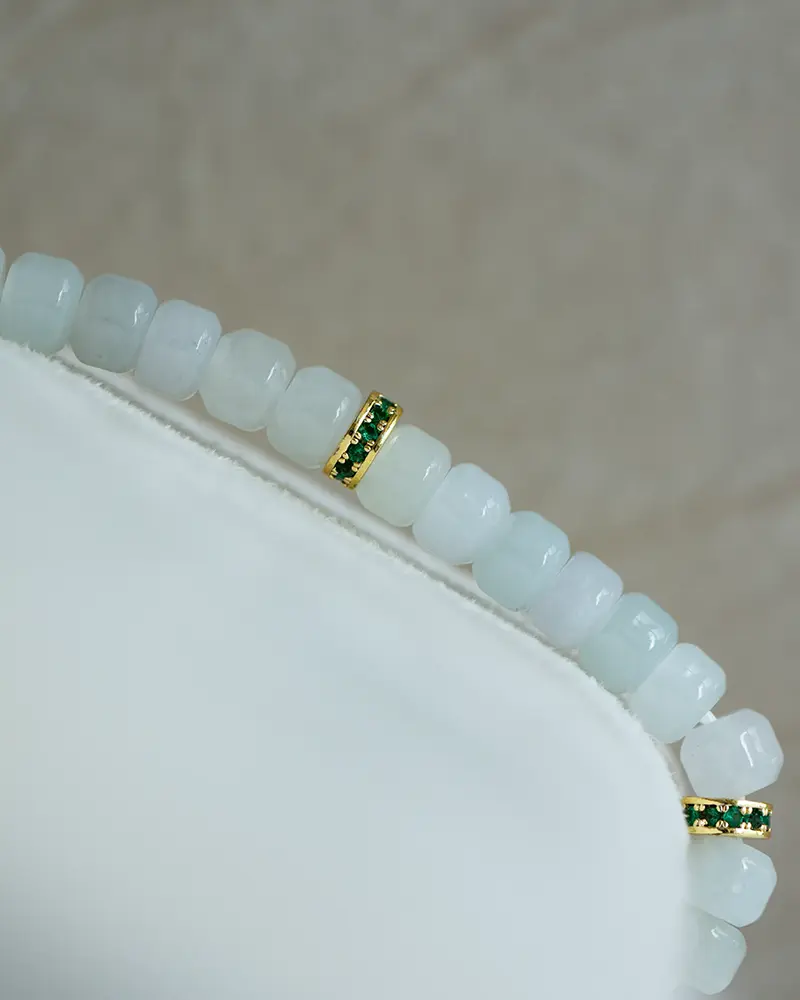 DSC_0734 拷贝 A close-up of a beaded bracelet featuring translucent white beads and two gold spacer beads accented with small green stones, displayed on a white surface with a neutral background.
