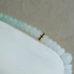 A close-up of a beaded bracelet with pale blue and white round beads and a single gold accent bead on a soft, light-colored surface.