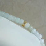 A close-up of a beaded bracelet with pale blue and white rectangular beads, featuring a single gold bead encrusted with small clear stones, displayed on a white surface with a neutral background.